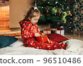 A little girl opens a Christmas present near the Christmas tree. Christmas time, gifts, festive mood. The concept of Christmas and New Year 96510486