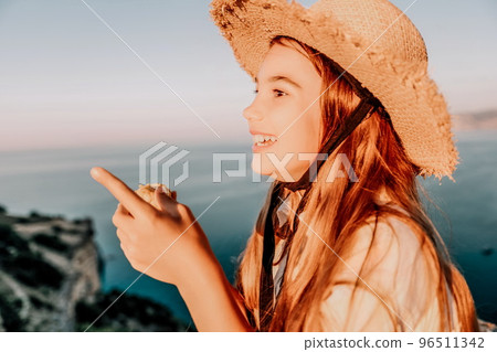 Happy girl eating corn. Summer snacking on the sea. Portrait of young beautiful girl in straw hat eating grilled corn while sitting by the sea on sunset time. Close up. Selective focus Happy girl eating corn. Summer snacking on the sea. Portrait of young beautiful girl in straw hat eating grilled corn while sitting by the sea on sunset time. Close up. Selective focus 96511342
