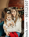 Beautiful mother and daughter on the background of a fireplace decorated for Christmas. Portrait of a loving family in close-up. Merry Christmas and Happy holidays 96512307