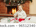 A baby girl drinks hot chocolate by the fireplace at home. A little girl on Christmas Eve or New Year's Eve at home. The concept of Christmas and New Year 96512340
