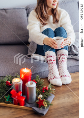 Woman in sweater and warm woolen socks on sofa holding cup of hot drink with cozy Christmas mood candles decor for hygge home on the coffee table. home coziness. Winter and Xmas holidays. Vertical. Woman in sweater and warm woolen socks on sofa holding cup of hot drink with cozy Christmas mood candles decor for hygge home on the coffee table. home coziness. Winter and Xmas holidays. Vertical. 96518948