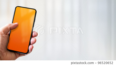 Hand holding a smartphone with blank orange screen. Office background. Horizontal banner. 96520652