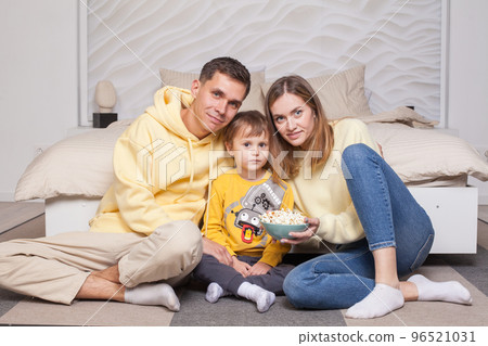 Friendly cute family, parents in yellow clothes with child son sitting by the bed and eating popcorn, looking to the camera Friendly cute family, parents in yellow clothes with child son sitting by the bed and eating popcorn, looking to the camera 96521031