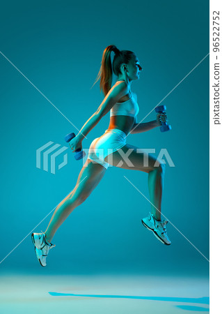 Young sportive girl in white sportswear training with dumbbells isolated over blue studio background in neon light. High jump 96522752