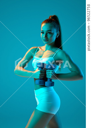 Young sportive girl in white sportswear training with dumbbells isolated over blue studio background in neon light Young sportive girl in white sportswear training with dumbbells isolated over blue studio background in neon light 96522758