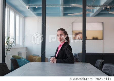 Successful young female leader in a suit with a pink shirt sitting in a modern glass office with a determined smile. Successful young female leader in a suit with a pink shirt sitting in a modern glass office with a determined smile. 96524115