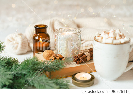 Hygge Cup of hot cocoa or coffee with marshmallow, sweater, candle, book, cinnamon and fir tree. Christmas lights cozy background. Xmas holiday wooden star decorations 96526114