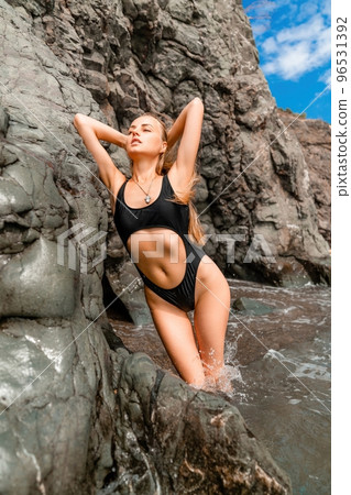 Woman swimsuit sea. Attractive blonde woman in a black swimsuit enjoying the sea air on the seashore around the rocks. Travel and vacation concept. 96531392