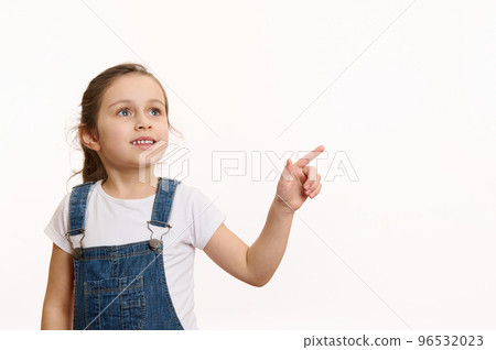 Horizontal studio shot on white background of a happy European little girl, adorable preschooler smart child, wearing blue denim overalls, pointing away on free space for your advertising content 96532023