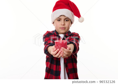 Adorable Caucasian preteen boy wearing Santa hat, holding a lit red candle to the camera, posing against a white background. Copy ad space. Merry Christmas and Happy New Year concept. Winter holidays 96532126