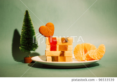 Marzipan marmalade turrone on plate, tangerine on green emerald background. Spanish dessert made from almonds, honey, sugar, eggs. Christmas sweets, heart-shaped cookie. New Year pine tree decor 2023. 96533539