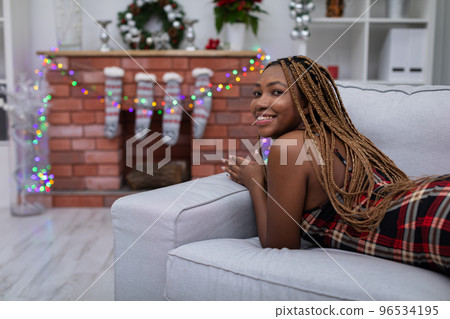 A dark-skinned girl lies on a sofa and looks smiling at the camera. Christmas decor. A dark-skinned girl lies on a sofa and looks smiling at the camera. Christmas decor. 96534195