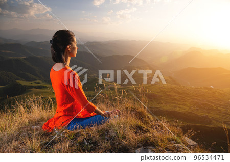 young caucasian woman meditates in the lotus position in the grass in the mountains near the cliff in the setting sun 96534471