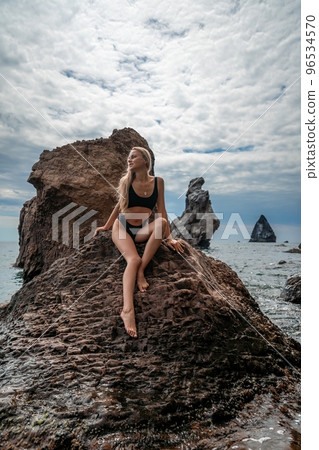 Woman swimsuit sea. Attractive blonde woman in a black swimsuit enjoying the sea air on the seashore around the rocks. Travel and vacation concept. 96534570