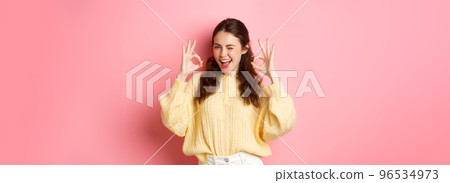 Portrait of cheerful pretty girl winking and saying yes, showing okay sign, alright gesture, praise good job, well done, standing pleased against pink background Portrait of cheerful pretty girl winking and saying yes, showing okay sign, alright gesture, praise good job, well done, standing pleased against pink background 96534973