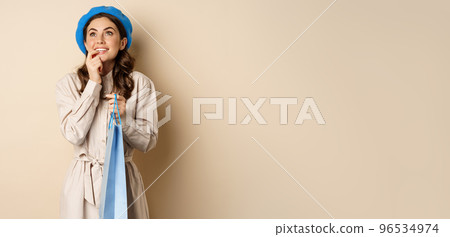 Image of modern beautiful woman posig with shopping gift bag, smiling and looking up thoughtful, thinking, standing over beige background Image of modern beautiful woman posig with shopping gift bag, smiling and looking up thoughtful, thinking, standing over beige background 96534974