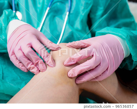 A traumatologist wearing medical gloves palpates the injured knee during a routine examination of the patient. A traumatologist wearing medical gloves palpates the injured knee during a routine examination of the patient. 96536857