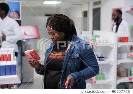 African american customer buying heart medication in drugstore, reading instruction, holding tablets package, standing in pharmacy aisle. Client choosing vitamins, medium shot African american customer buying heart medication in drugstore, reading instruction, holding tablets package, standing in pharmacy aisle. Client choosing vitamins, medium shot 96537766