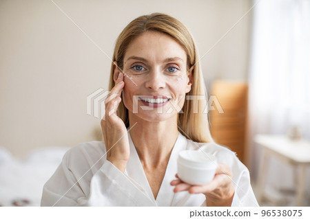 Beautiful middle aged woman in silky bathrobe using nourishing eye cream, enjoying face care routine Beautiful middle aged woman in silky bathrobe using nourishing eye cream, enjoying face care routine 96538075