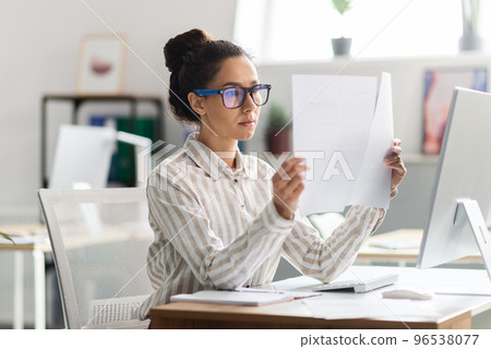 Serious busy woman in glasses sitting at table with computer and working with documents in office interior Serious busy woman in glasses sitting at table with computer and working with documents in office interior 96538077