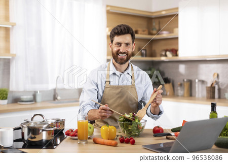 Smiling adult european bearded male chef in apron preparing salad in modern kitchen interior Smiling adult european bearded male chef in apron preparing salad in modern kitchen interior 96538086