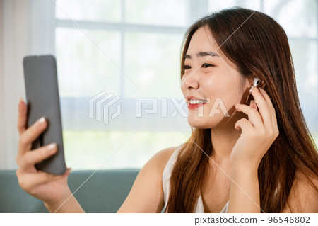 Lifestyle Asian young woman wearing wireless earphones listening to favorite song on her smart mobile phone, Happy relaxed female wear earbuds listen song on smartphone in bright living room at home 96546802