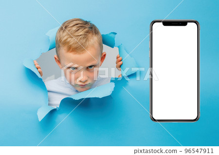 Angry cute boy is having fun played on blue background wall, climbs through a hole in the paper near huge mobile phone. Angry cute boy is having fun played on blue background wall, climbs through a hole in the paper near huge mobile phone. 96547911