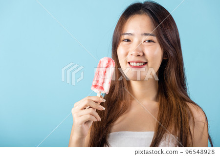 Sweet tasty frozen dessert on summer time. Portrait of happy Asian young beautiful woman smiling and holding delicious ice cream wood stick mixed fruit flavor, studio shot isolated on blue background 96548928