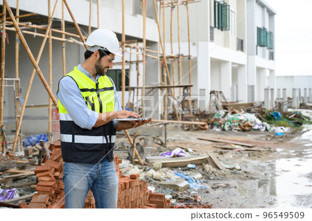 Young attractive construction smiling bearded in vest with white helmet working with laptop, standing on building construction site. Home building project. Young attractive construction smiling bearded in vest with white helmet working with laptop, standing on building construction site. Home building project. 96549509