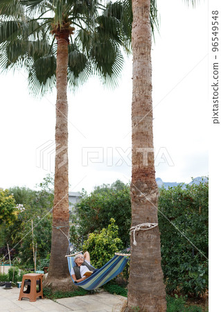 Smiling senior good-looking grey hair woman wearing glasses while reading in hammock in the summer garden under palms Smiling senior good-looking grey hair woman wearing glasses while reading in hammock in the summer garden under palms 96549548