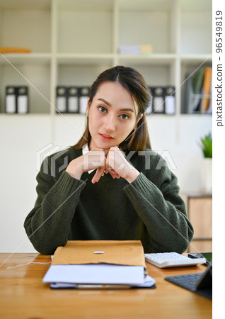 Charming Asian female hands on chin, smiling and looking at camera while sitting at her desk. Charming Asian female hands on chin, smiling and looking at camera while sitting at her desk. 96549819