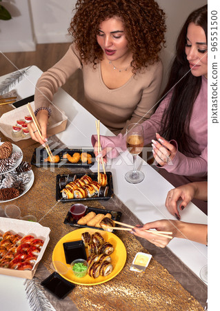 Portrait of three beautiful young women eating japanese food and drinking wine at home. Portrait of three beautiful young women eating japanese food and drinking wine at home. 96551500