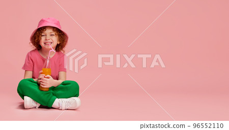 Portrait of cute little girl, child with curly red hair sitting in panama and drinking juice isolated over pink background. Flyer Portrait of cute little girl, child with curly red hair sitting in panama and drinking juice isolated over pink background. Flyer 96552110