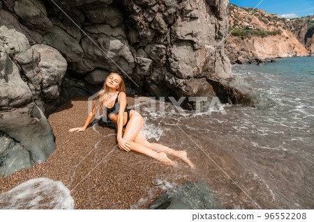 Woman swimsuit sea. Attractive blonde woman in a black swimsuit enjoying the sea air on the seashore around the rocks. Travel and vacation concept. 96552208