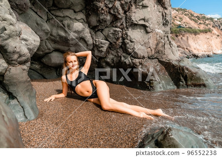 Woman swimsuit sea. Attractive blonde woman in a black swimsuit enjoying the sea air on the seashore around the rocks. Travel and vacation concept. 96552238
