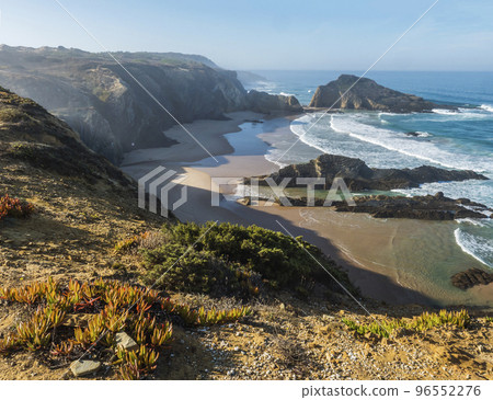 View of empty Praia da Zambujeira do Mar beach with ocean waves, cliffs and stones, wet golden sand and green vegetation at wild Rota Vicentina coast, Odemira, Portugal. 96552276