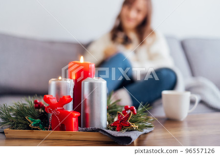 Christmas cozy mood composition for hygge home decor. Burning candles, pine tree branches on tray on the coffee table with resting woman drinking hot tea and petting cat on the sofa. Selective focus. 96557126