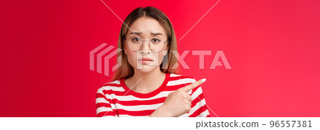 Sad cute asian female coworker tired working wear glasses frowning, pointing left look camera upset drained feel pressured distressed, stand red background unhappy wear striped t-shirt 96557381