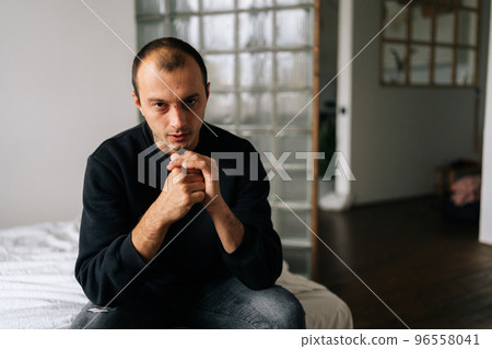 Portrait of serious young man sitting alone at home and looking at camera with serious expression holding hands on chin, deep thoughts feeling lonely. Frustrated male pondering make difficult choice. Portrait of serious young man sitting alone at home and looking at camera with serious expression holding hands on chin, deep thoughts feeling lonely. Frustrated male pondering make difficult choice. 96558041