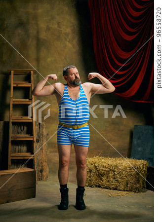 Strength. Mature man, retro circus strongman wearing striped sports swimsuit posing isolated over dark vintage circus backstage background. Art, 30s, 40s fashion style Strength. Mature man, retro circus strongman wearing striped sports swimsuit posing isolated over dark vintage circus backstage background. Art, 30s, 40s fashion style 96558720