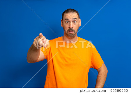 Latino man with a beard wearing an orange t-shirt pointing forward with a surprised expression. 96560960