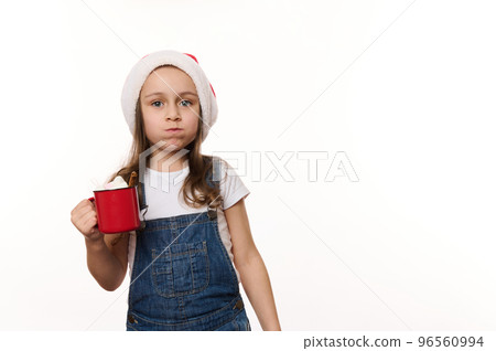 Mischievous 5 years old little girl wearing Santa hat and denim overalls, drinking hot chocolate drink and eating marshmallows, isolated on white background with copy ad space. Christmas atmosphere 96560994