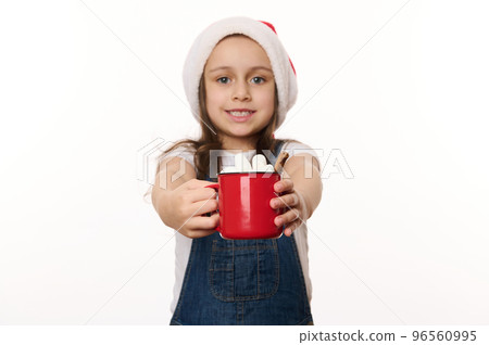 Selective focus on a red cup of hot cocoa drink with marshmallows, in the hands of a blurred lovely baby girl wearing Santa hat. Traditional food and beverage for Christmas. Winter festive atmosphere Selective focus on a red cup of hot cocoa drink with marshmallows, in the hands of a blurred lovely baby girl wearing Santa hat. Traditional food and beverage for Christmas. Winter festive atmosphere 96560995