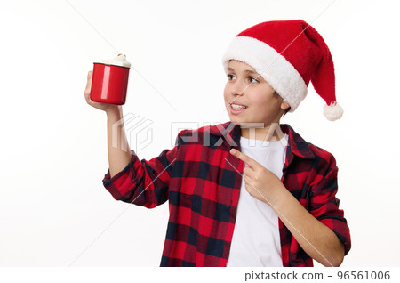Handsome multi-ethnic preteen boy in Santa hat, pointing on red cup of hot cocoa with marshmallows, isolated on white background. Copy space or promotional text. Christmastime. Happy winter holidays Handsome multi-ethnic preteen boy in Santa hat, pointing on red cup of hot cocoa with marshmallows, isolated on white background. Copy space or promotional text. Christmastime. Happy winter holidays 96561006