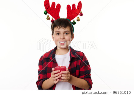 Happy Caucasian teenage boy with deer antler hoop on his head, smiling a beautiful toothy smile, holding a lit Christmas candle, looking at camera. Isolated on white background. Copy advertising space 96561010
