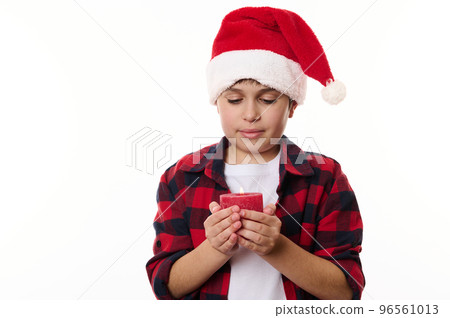 Close-up portrait of handsome Caucasian teenage boy wearing Santa hat and red plaid shirt, making a cherished wish while holding a lit Christmas candle. Isolated on white background. Copy ad space Close-up portrait of handsome Caucasian teenage boy wearing Santa hat and red plaid shirt, making a cherished wish while holding a lit Christmas candle. Isolated on white background. Copy ad space 96561013