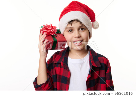 Adorable kid boy in Santa hat, shakes Christmas gift box, guessing the present inside it, smiling a beautiful smile looking at camera, isolated close-up portrait on white background with copy space 96561016