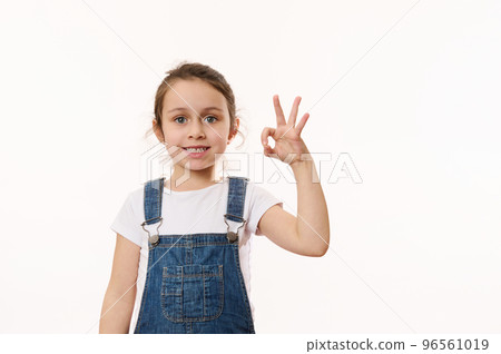 Adorable elementary age, preschool little child girl, in blue denim overalls, gestures, shows peace sign, cutely smiling cheerful toothy smile, looking at camera, isolated white background. Copy space 96561019