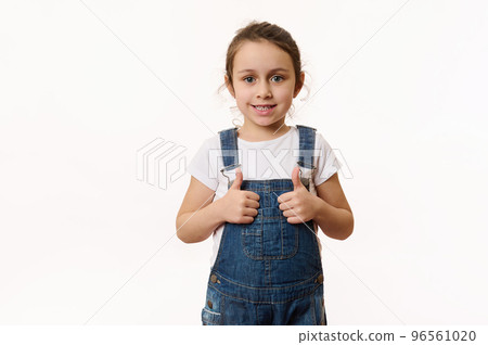 Adorable elementary age Caucasian little child, preschool kid, a lovely baby girl wearing blue denim overalls, shows thumbs up and sweetly smiles looking at camera, isolated on white background 96561020
