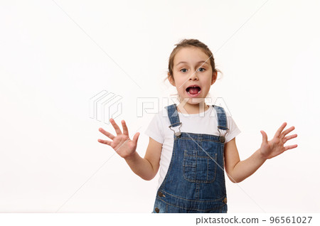 Emotional portrait on white background of a preschool child, overjoyed cute baby girl, wearing blue denim overalls, sweetly smiling, showing her hands palms to camera. Beautiful kids, happy childhood 96561027